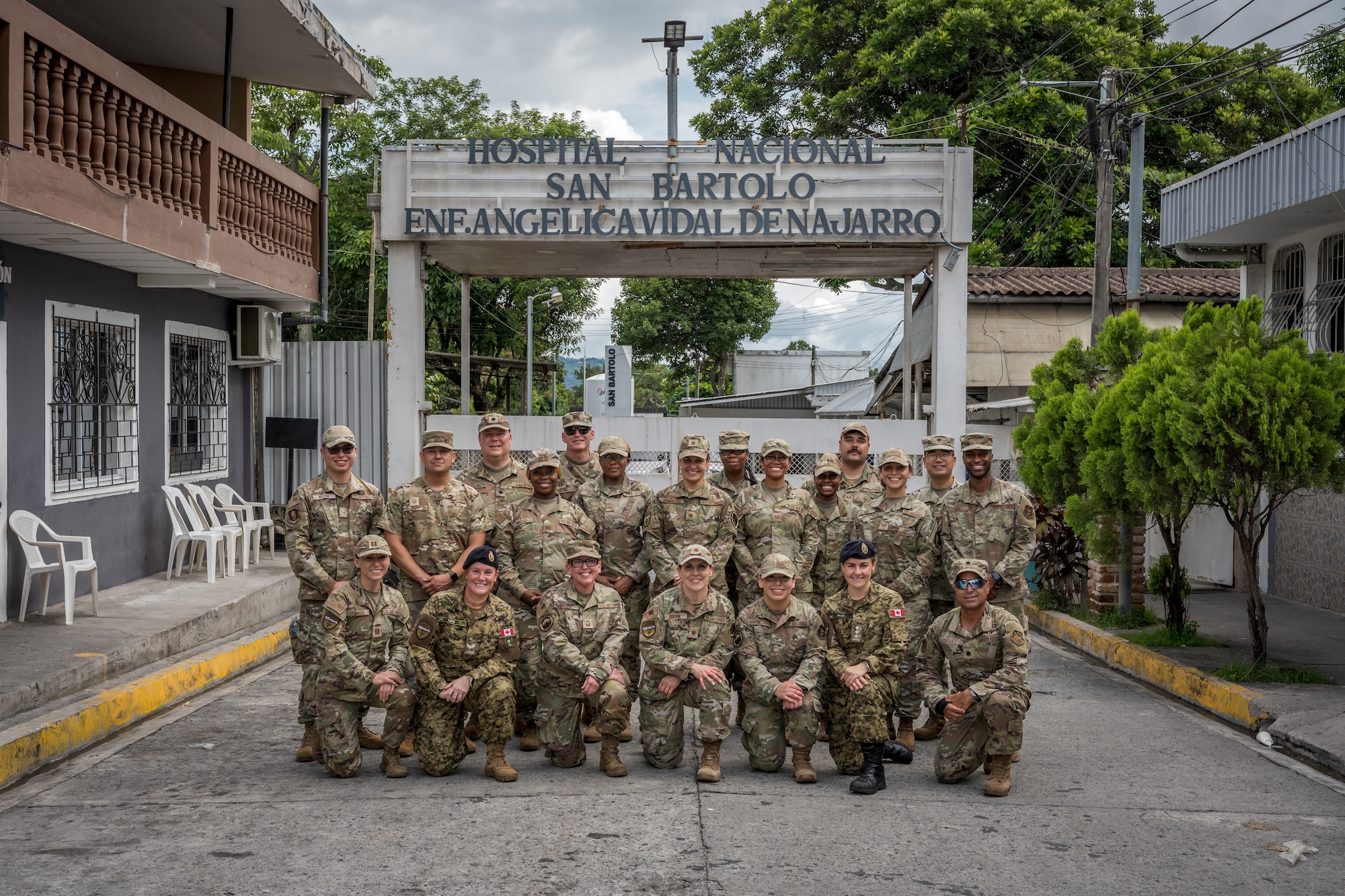 A group of international forces pose for a group photo in front of a sign that reads "HOSPITAL NACIONAL SAN BARTOLO ENF. ANGELICA VIDAL DENAJARRO"