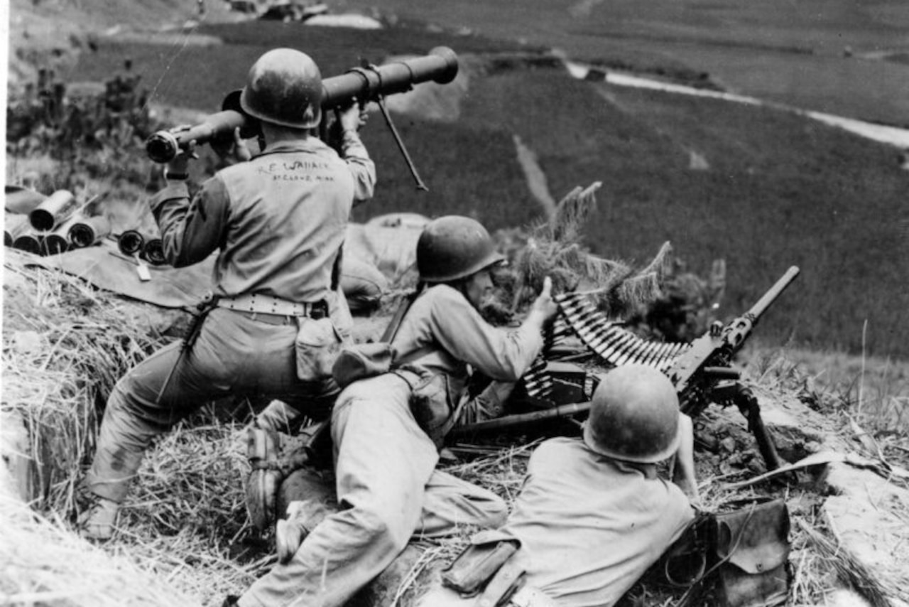 A soldier feeds ammunition into a machine gun as another man operates it. A third man prepares to fire a bazooka.