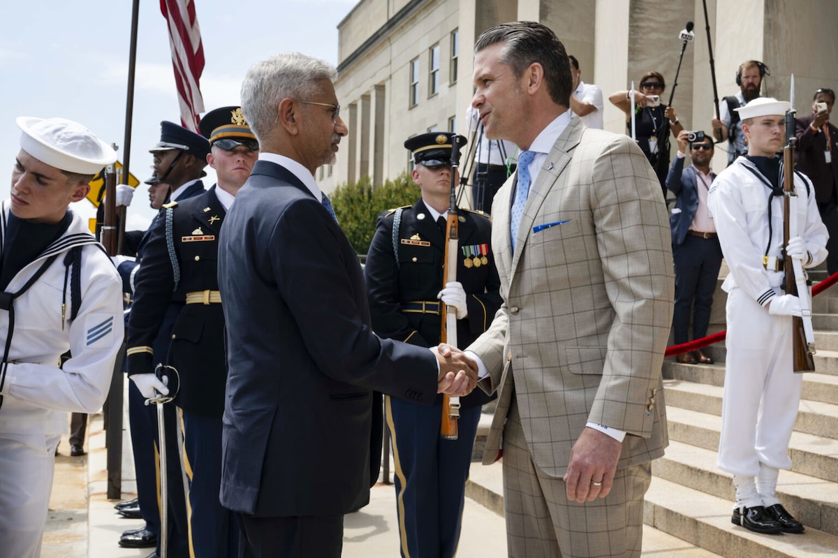 Two men wearing business attire shake hands while standing outdoors. Uniformed military personnel stand nearby with flags and ceremonial rifles.