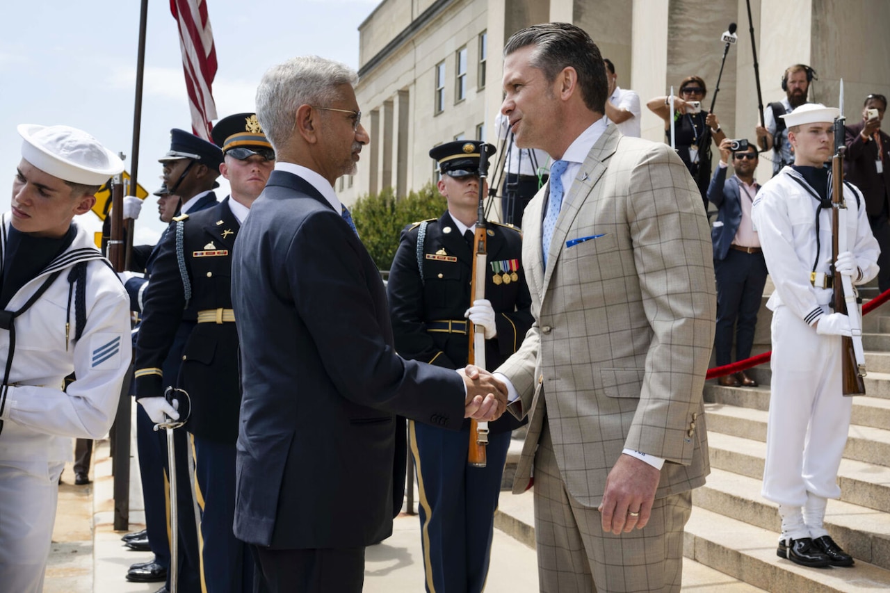 Two men wearing business attire shake hands while standing outdoors. Uniformed military personnel stand nearby with flags and ceremonial rifles.