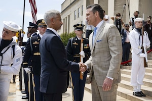 Two men wearing business attire shake hands while standing outdoors. Uniformed military personnel stand nearby with flags and ceremonial rifles.
