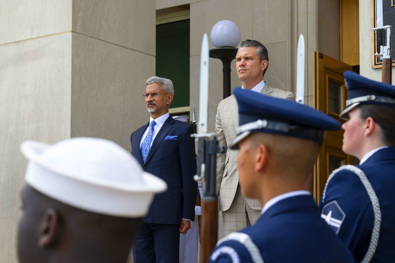 Two men wearing business attire stand next to each other. Uniformed military personnel are lined up near them with ceremonial rifles.