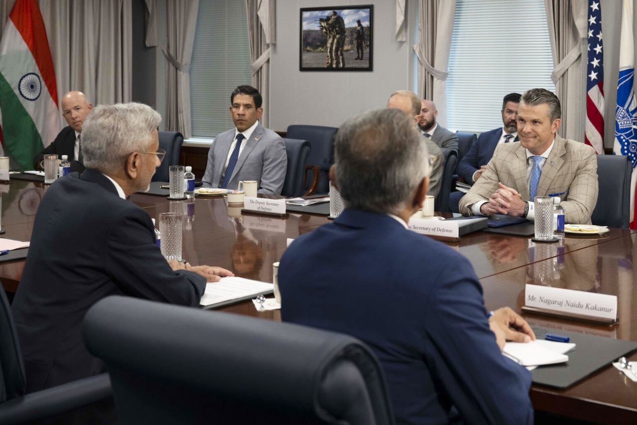 Men wearing business attire sit across from each other at a table. Behind them are an American flag, the flag of India and a U.S. Army flag. There are name tags on the table for each man.