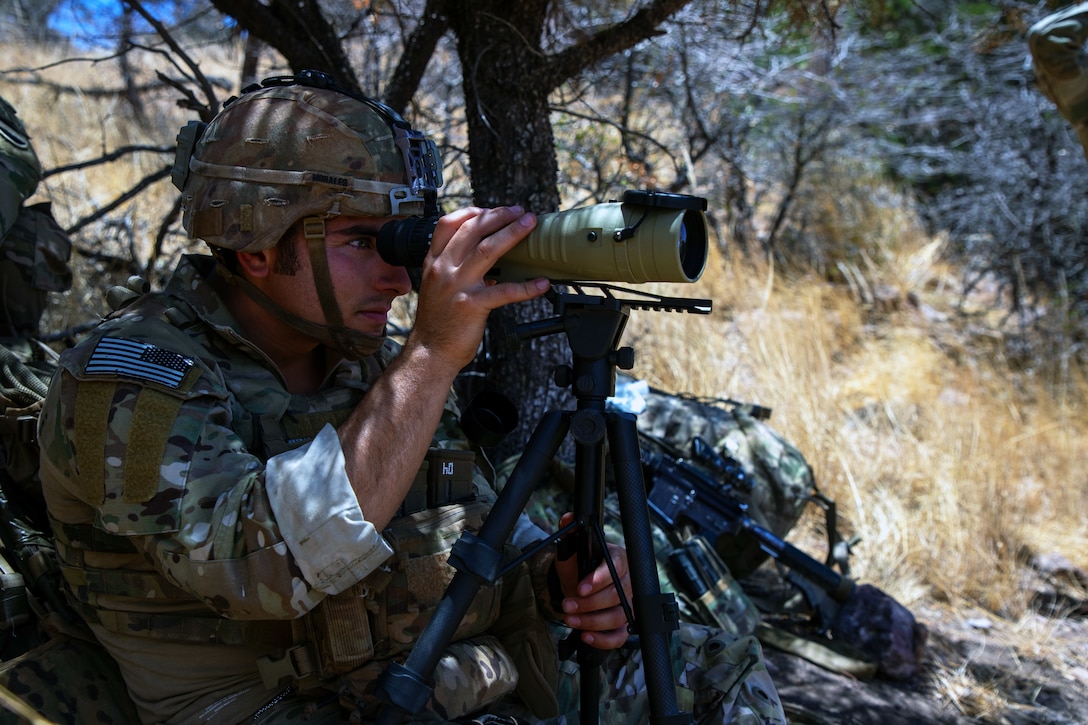A soldier looks through a small telescope under the cover of trees on a sunny day.