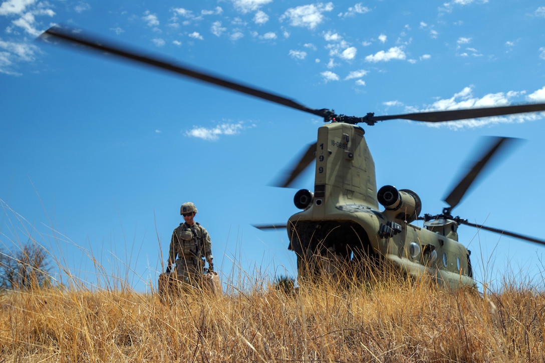 A soldier carrying fuel containers in each hand walks away from a helicopter on a field with sparse vegetation.