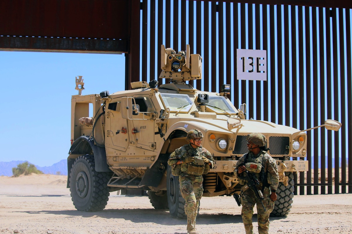Two soldiers in combat gear walk away from a military vehicle on a sunny day in desert-like conditions, with a tall security barrier in the background.