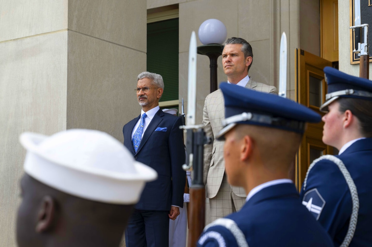 Two men wearing business attire stand next to each other. Uniformed military personnel are lined up near them with ceremonial rifles.