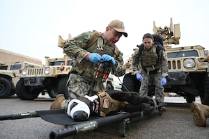 Two U.S. service members kneel on the ground over a canine casualty dummy on a litter in front of military Humvees during a large-scale joint medical exercise.