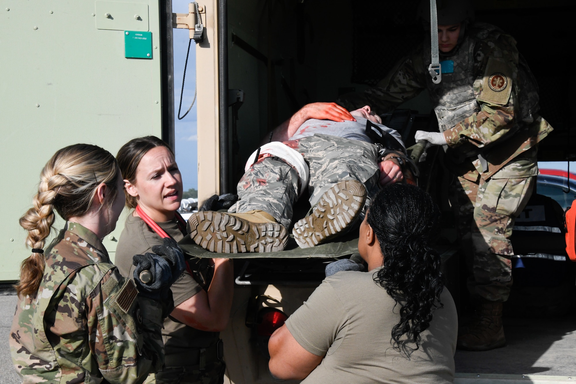 Three female Airmen hoist a wounded Airman on a litter into the back of an ambulance.