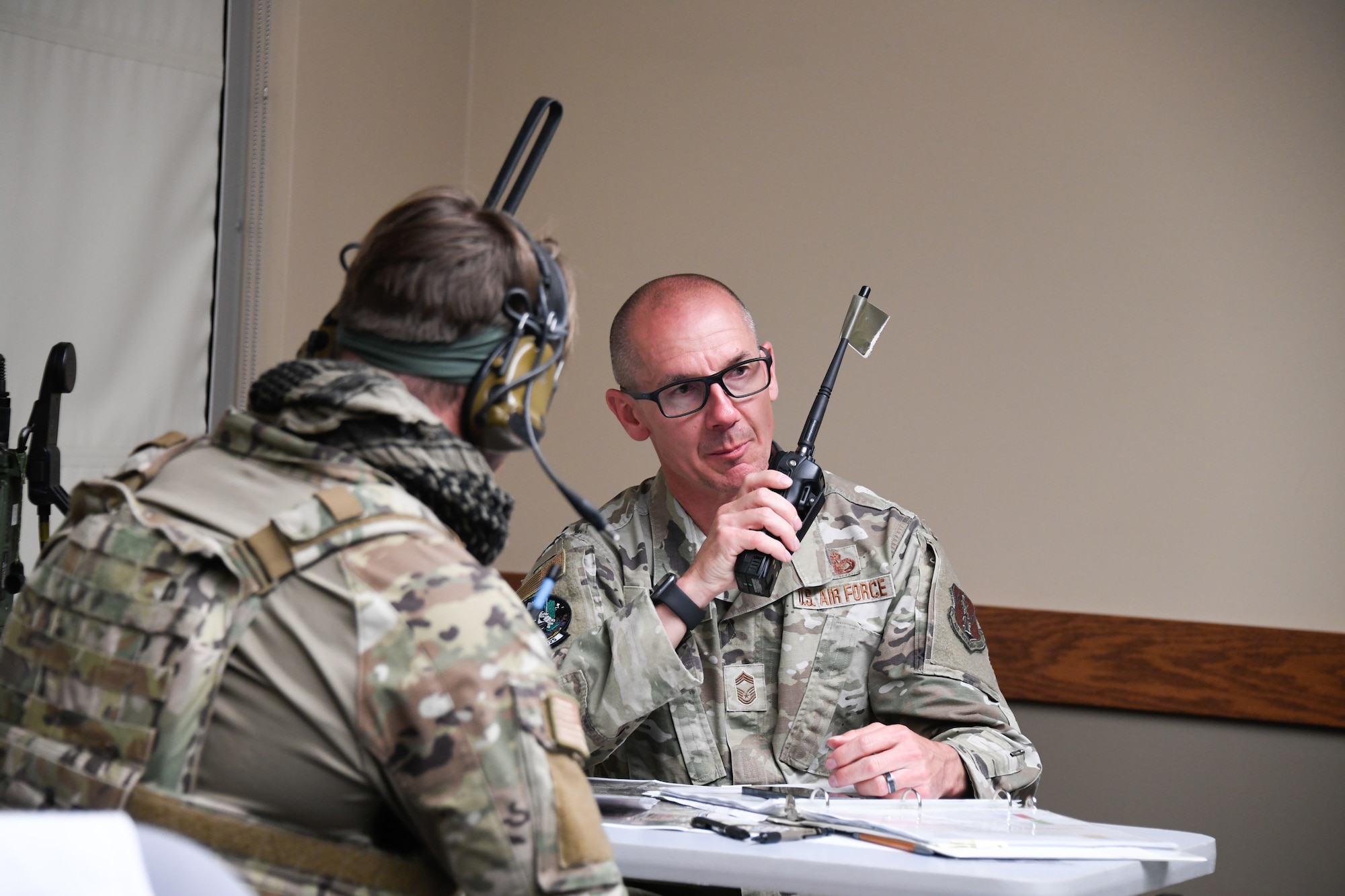 An Airman is seen over the shoulder of another Airman holding a radio to his mouth while seated at a table.