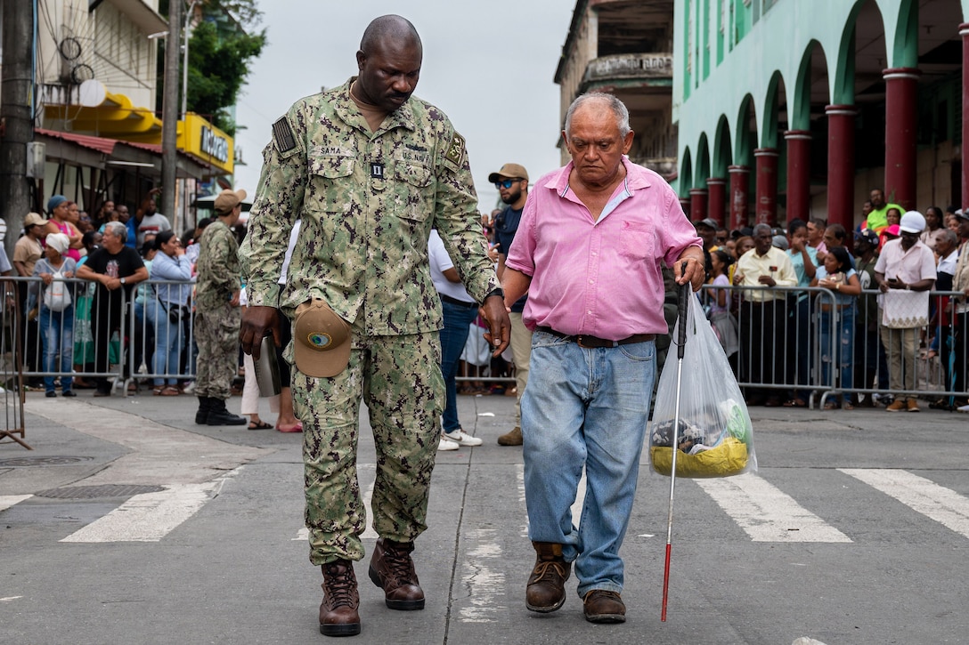 A sailor guides a person using a cane along a road in an urban area with a group of people standing behind gates in the background.
