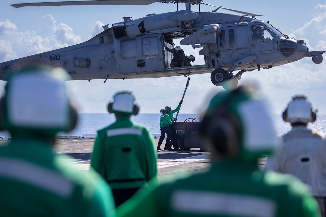 Two sailors on the deck of a ship hook a long device to the underside of a hovering helicopter as other sailors look on in the foreground.