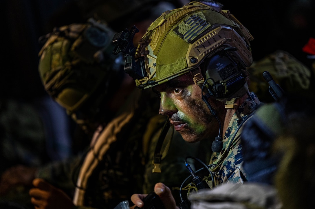 A Marine in combat gear wearing green and black face paint stares intently downward; another person in combat gear is next to the Marine.