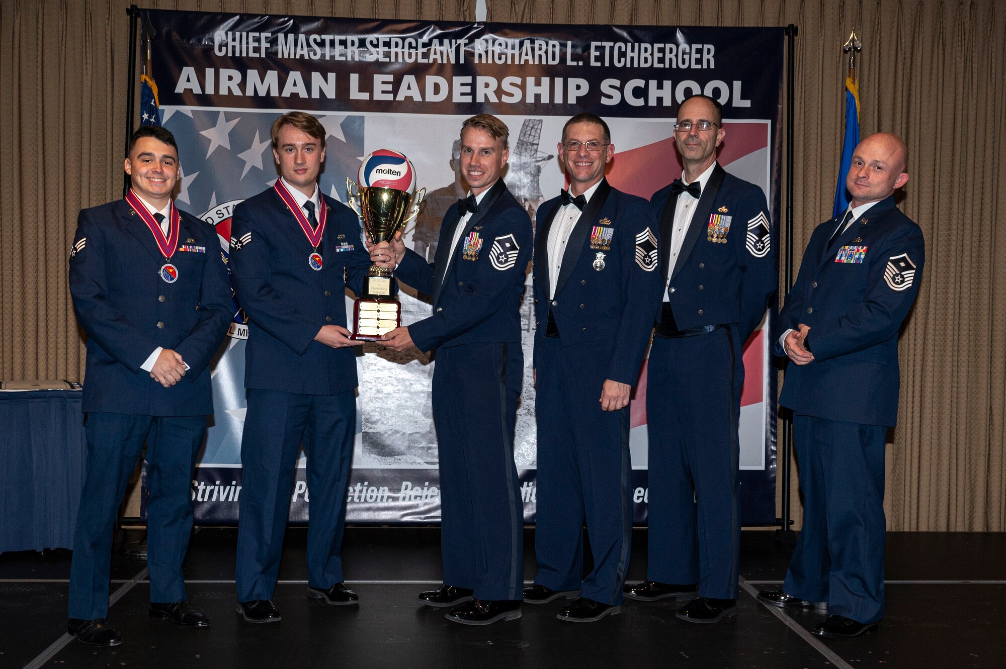 A group of six men stand on a stage while hold a trophy between the second and third men.