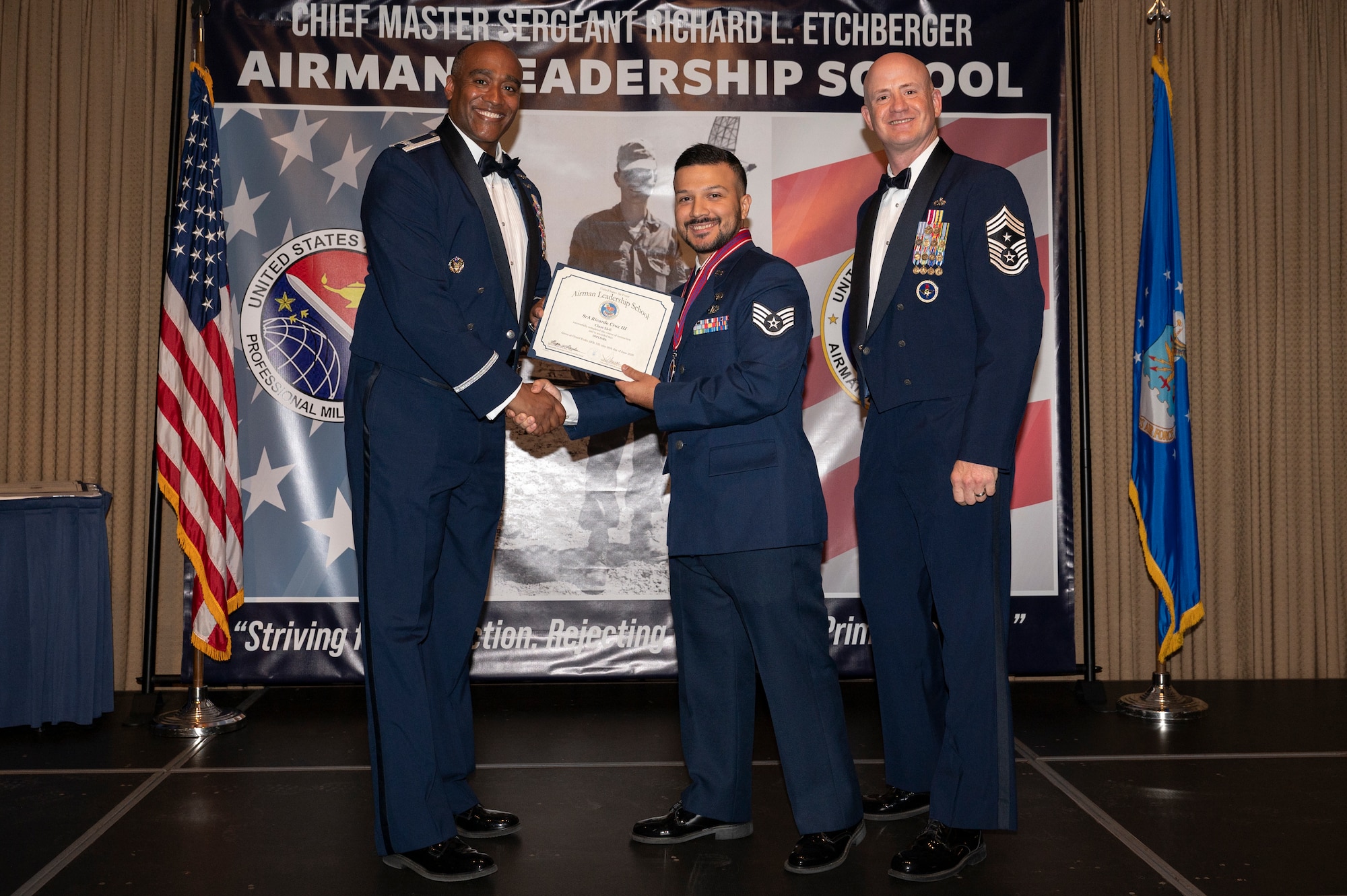 Three men stand on a stage together; the first man shake hands with the second man while holding a certificate together.