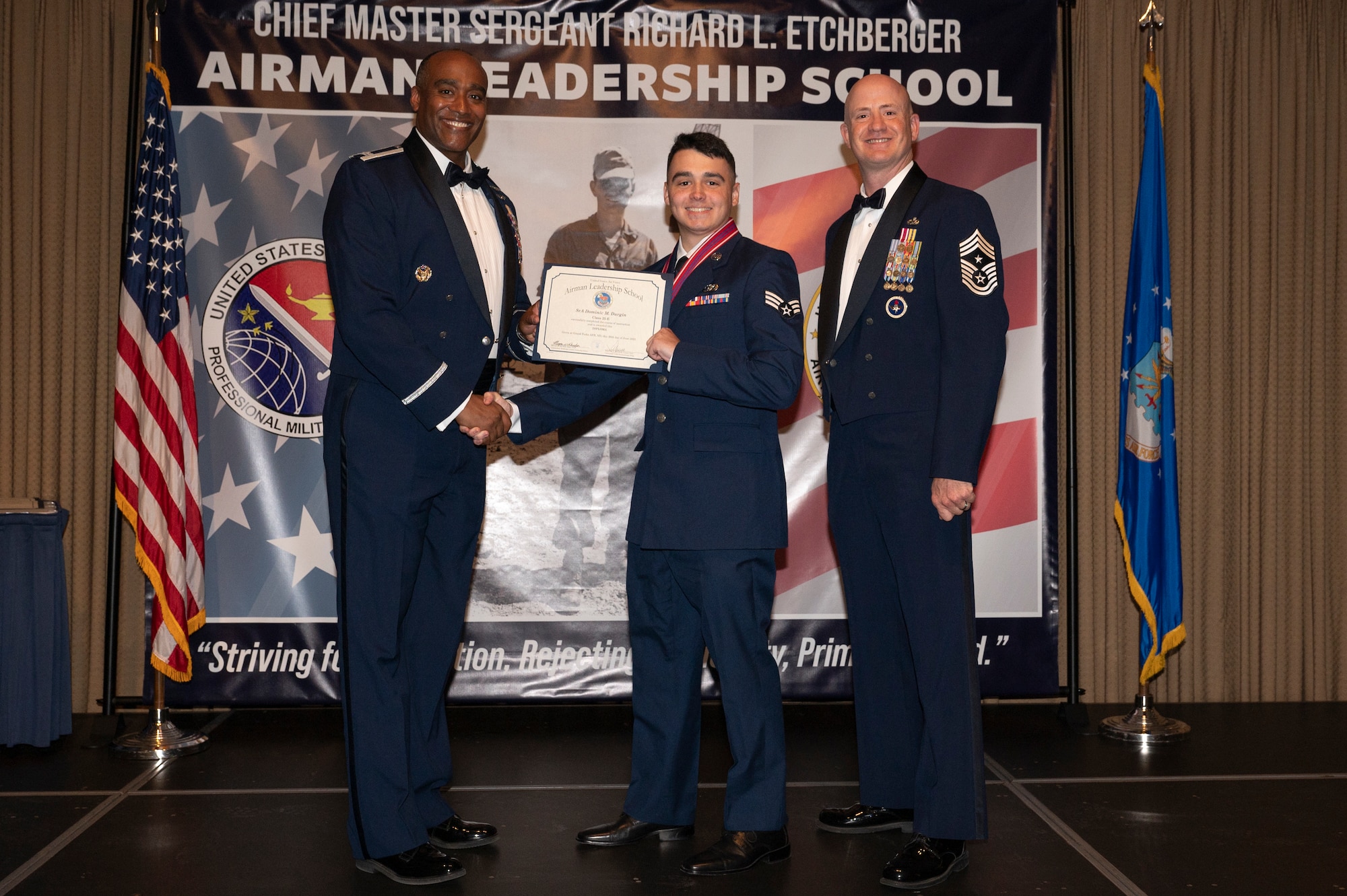 Three men stand on a stage together; the first man shake hands with the second man while holding a certificate together.