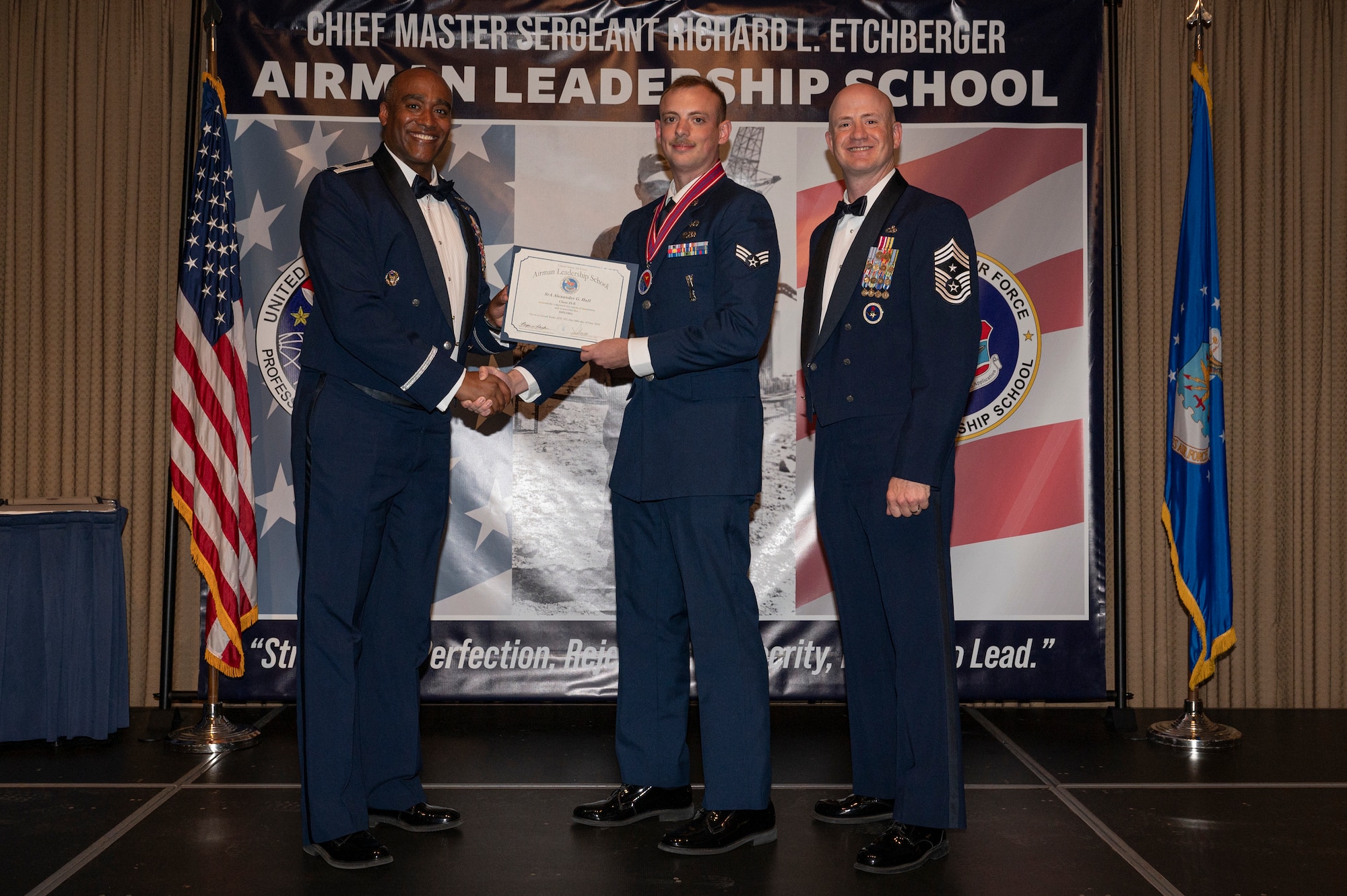 Three men stand on a stage together; the first man shake hands with the second man while holding a certificate together.