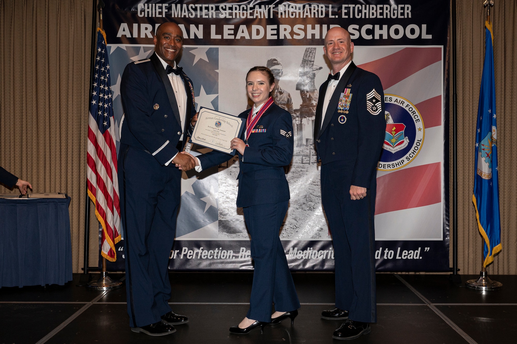 Two men stand on a stage with a woman standing between them, the first man shake hands with the woman while holding a certificate together.