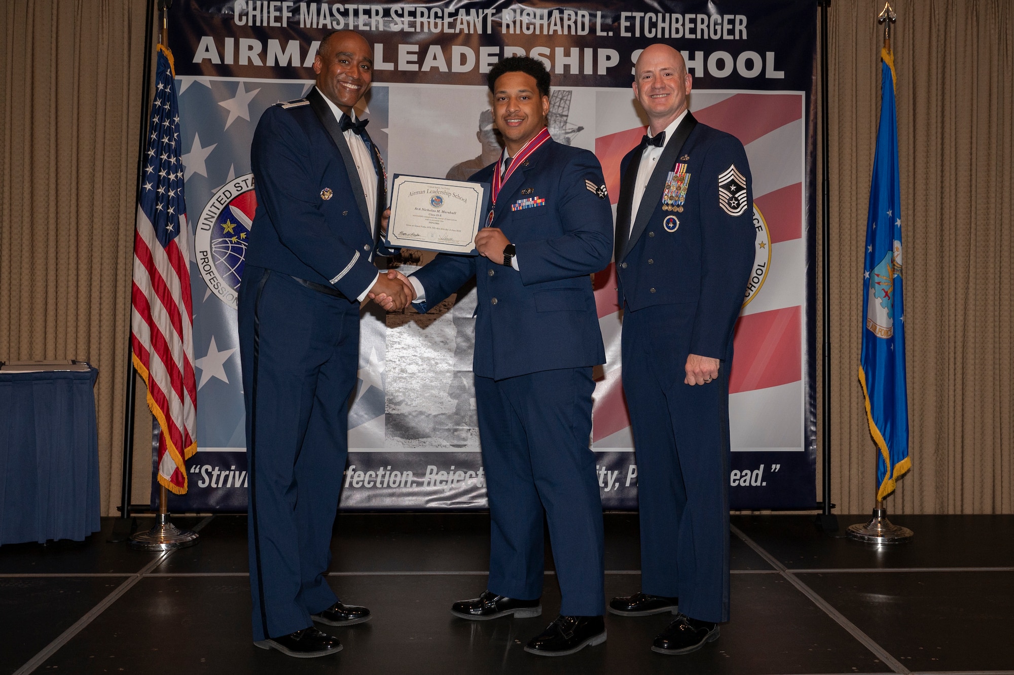 Three men stand on a stage, the first and second men shake hands while holding a certificate together.