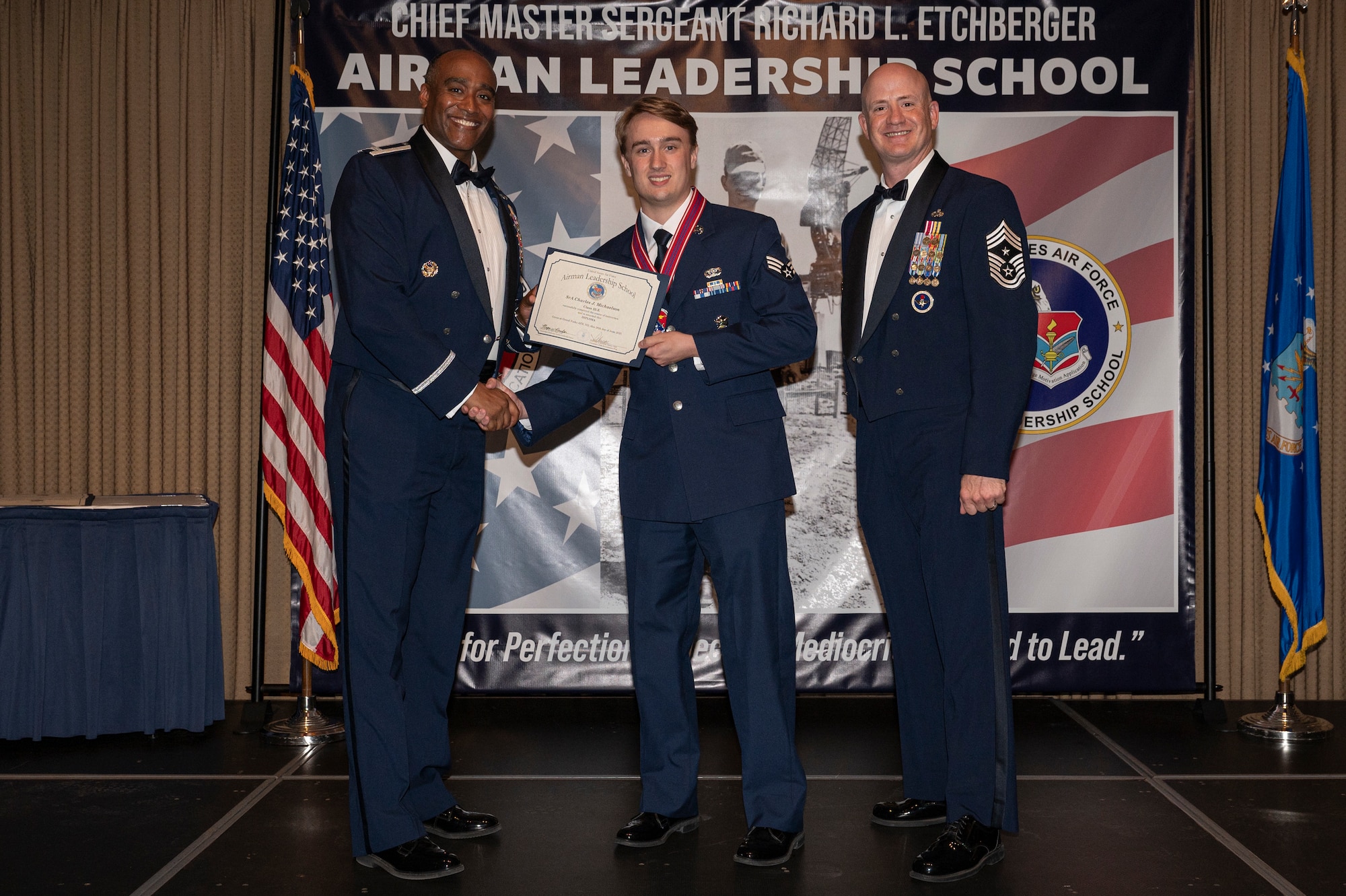Three men stand on a stage, the first and second men shake hands while holding a certificate together.