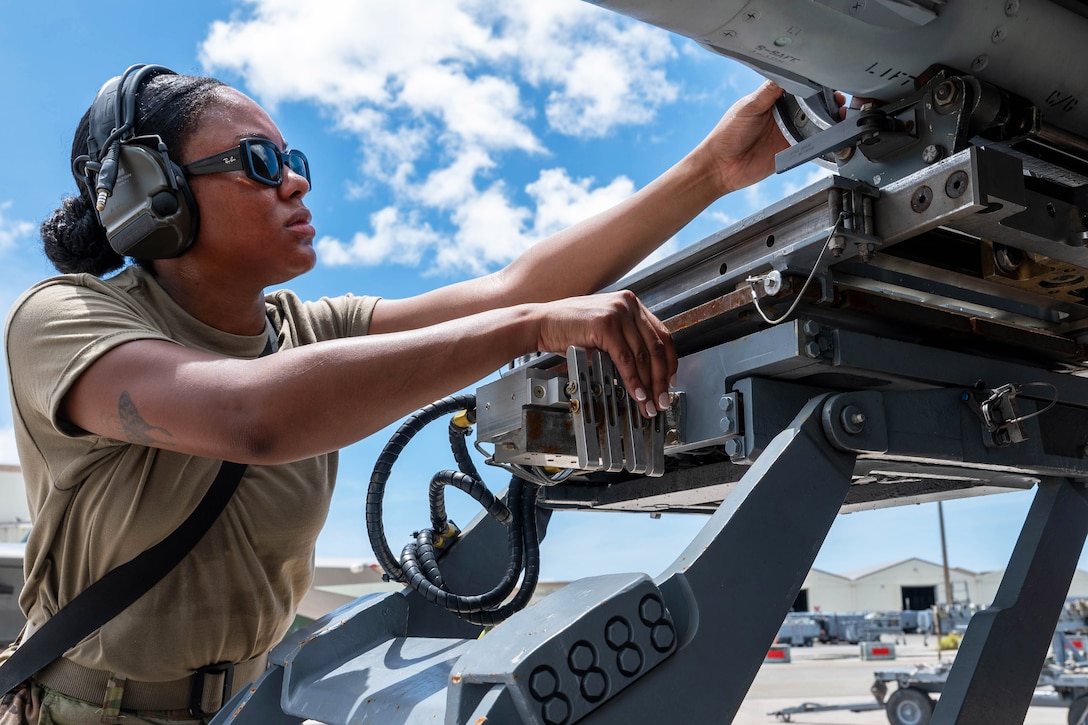 An airman wearing ear protection and sunglasses uses her hands to secure a missile to the underside of an aircraft parked on a runway.