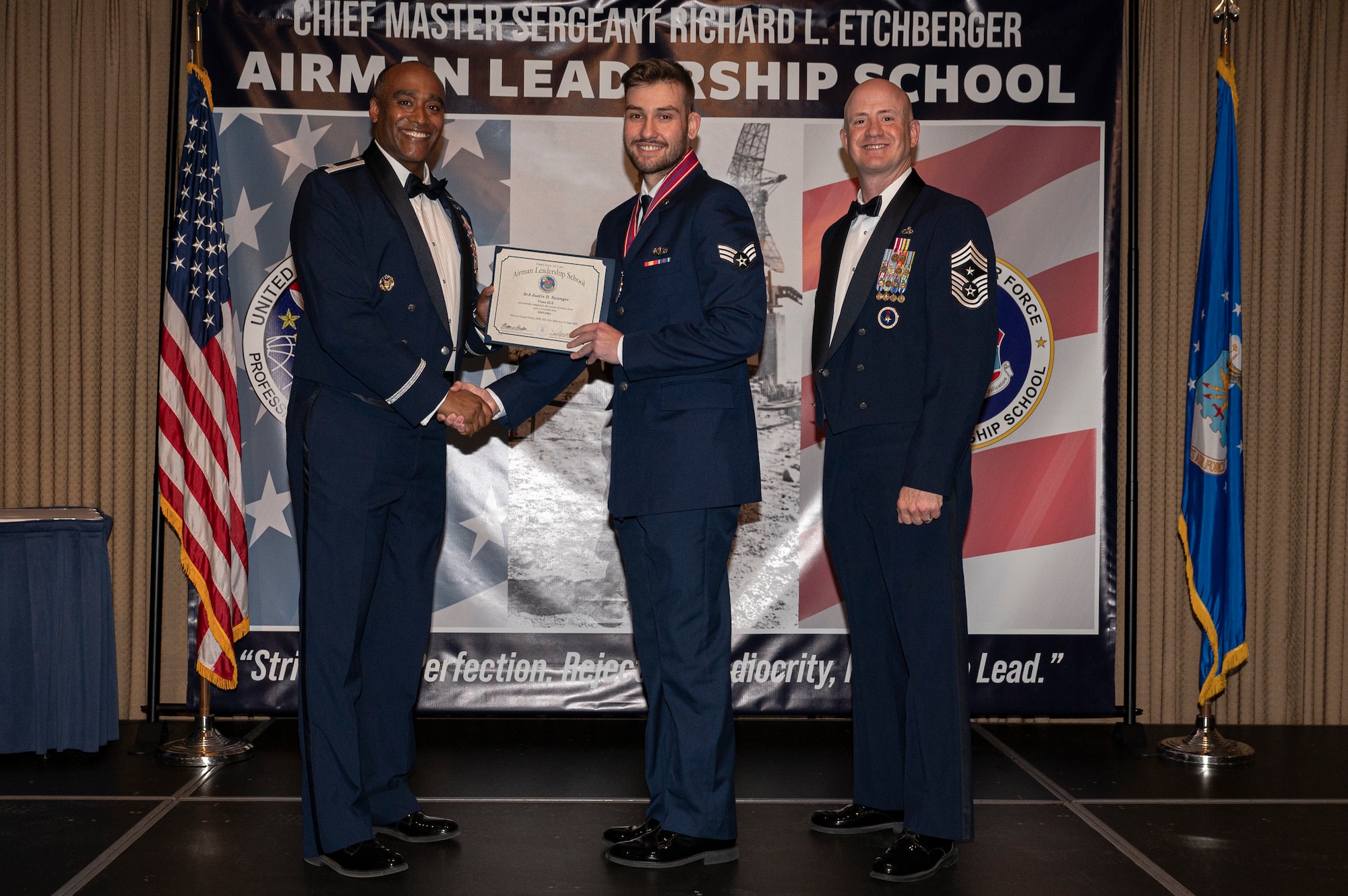 Three men stand on a stage, the first and second men shake hands while holding a certificate together.