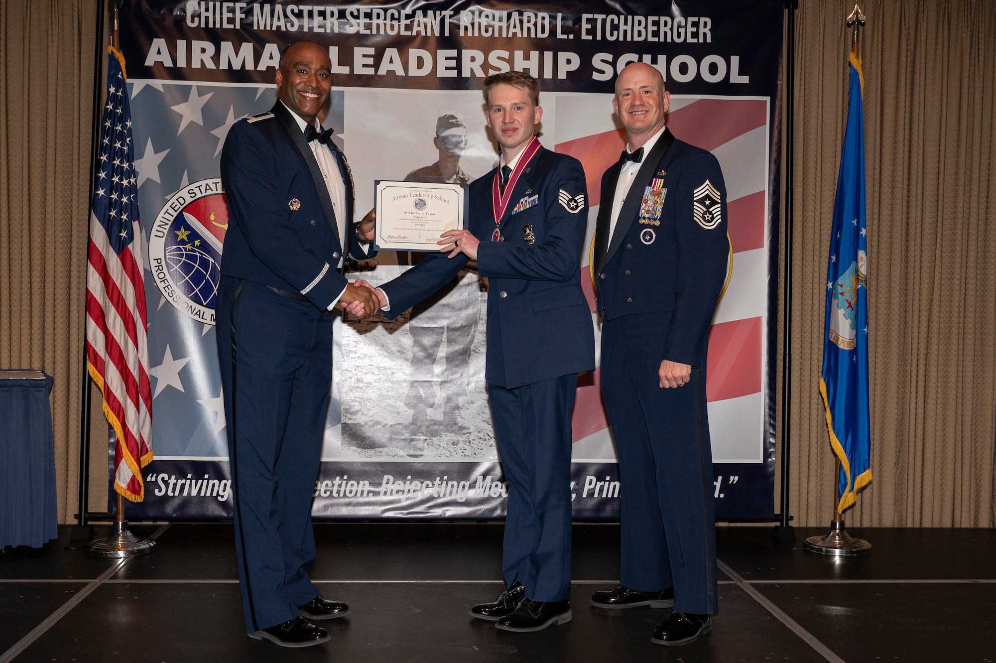 Three men stand on a stage, the first and second men shake hands while holding a certificate together.