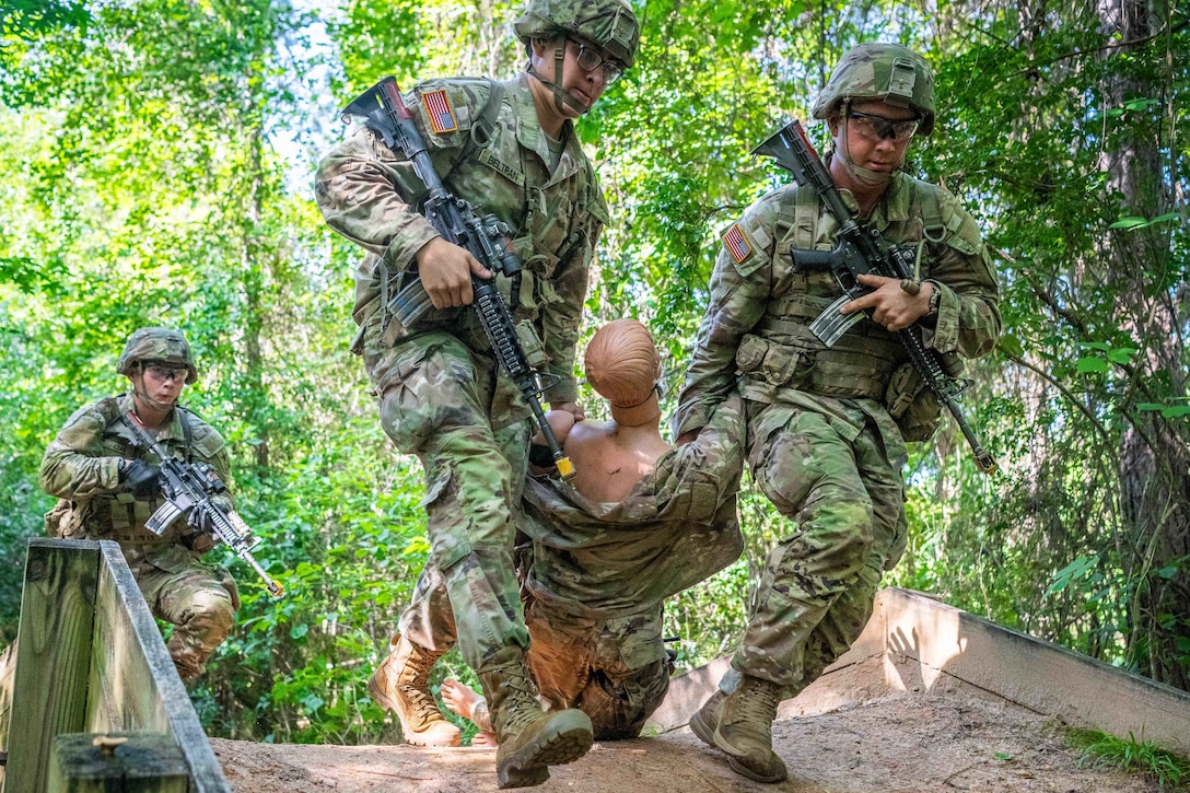 Two soldiers in combat gear pull a mannequin by the arms over a makeshift bridge in a wooded area as another soldier runs behind them.