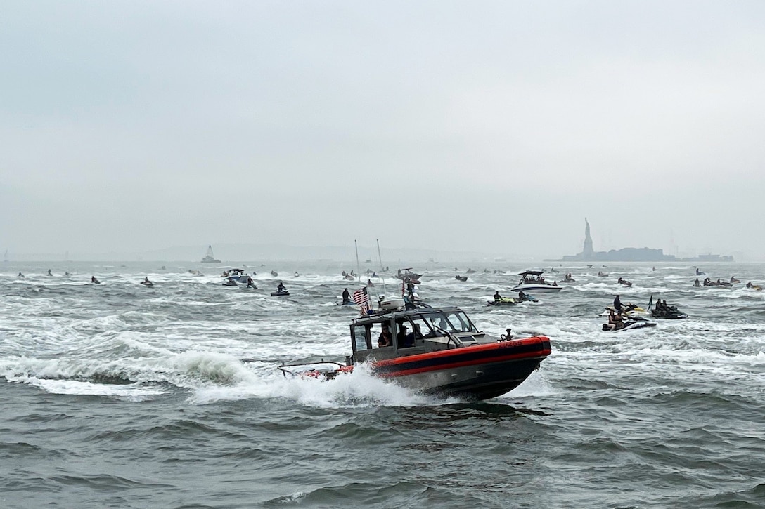 A Coast Guard vessel cruises alongside multiple Jet Skis and small boats in a river on a cloudy day, with the Statue of Liberty in the distance.