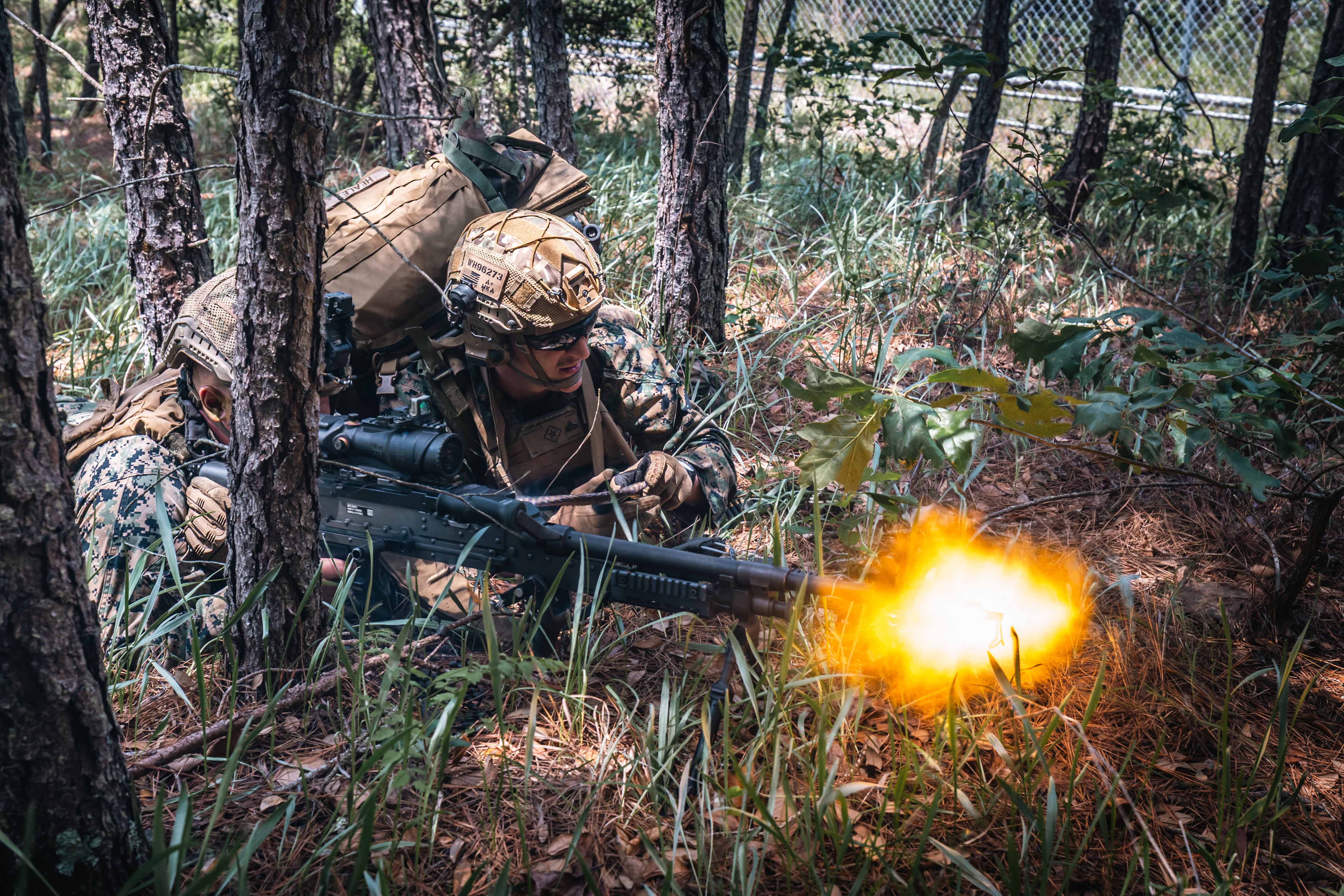 Two Marines lie in a wooded area as one fires a machine gun and the other looks toward the target. 