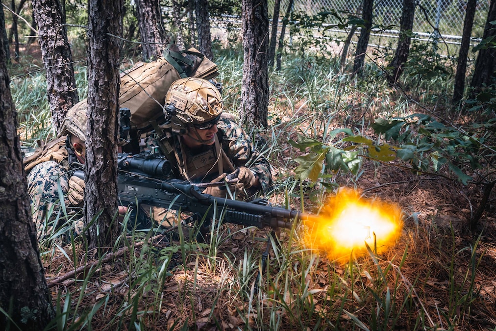 Two Marines lie in a wooded area as one fires a machine gun and the other looks toward the target.