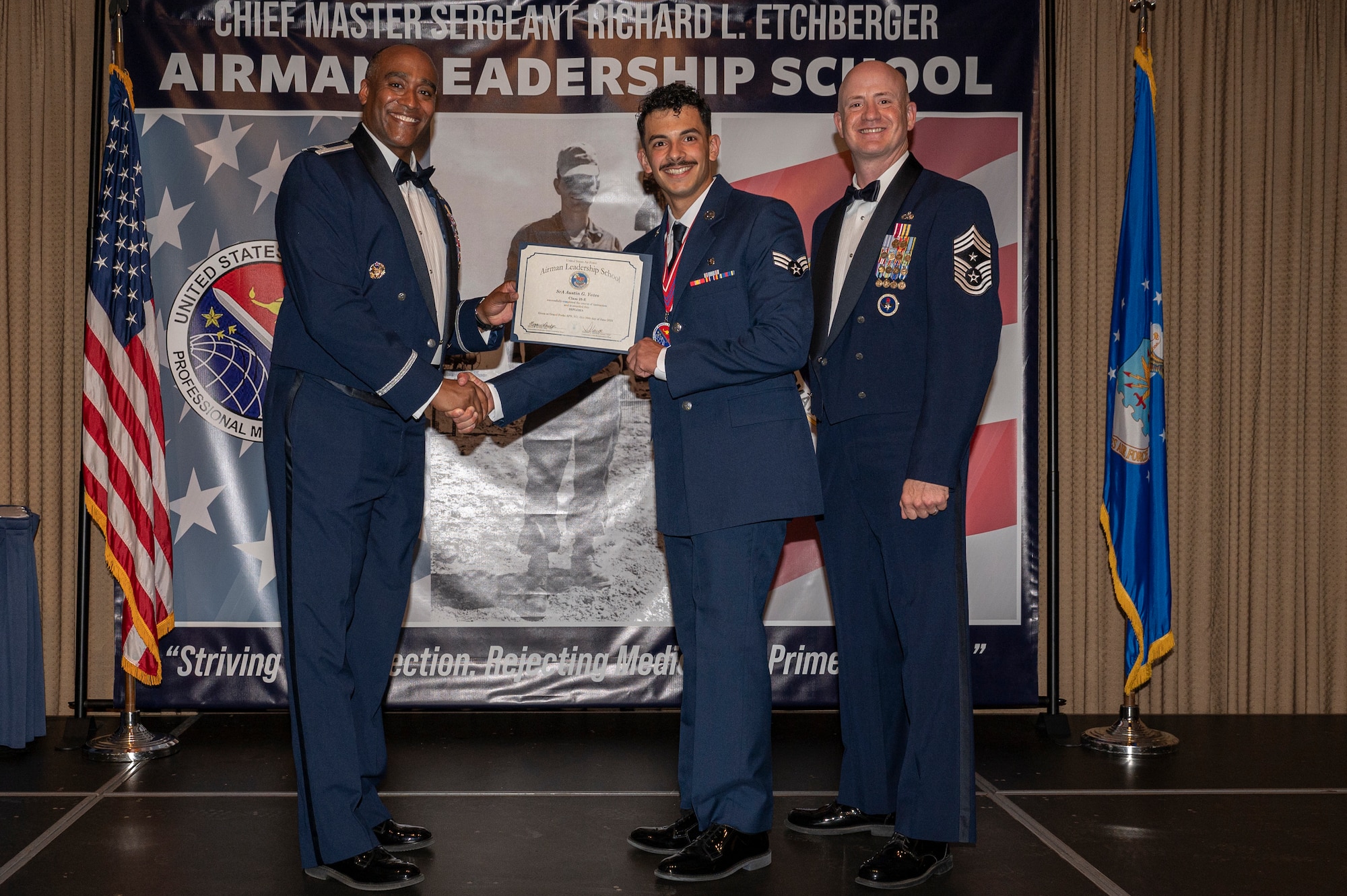 Three men stand on a stage together, while the first and second man shake hands while holding a certificate between them.