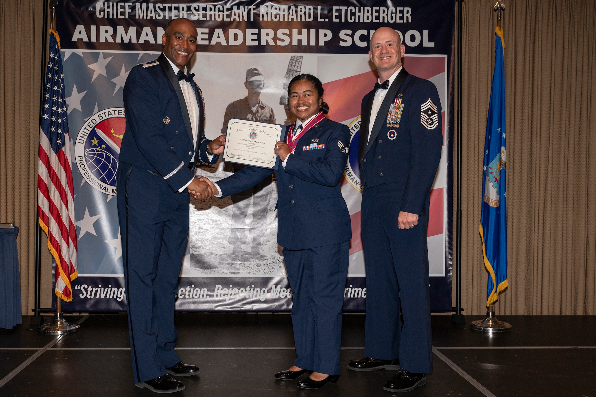 Two men stand on a stage with a woman standing between them while the first man shake hands with the woman, while holding an award together.