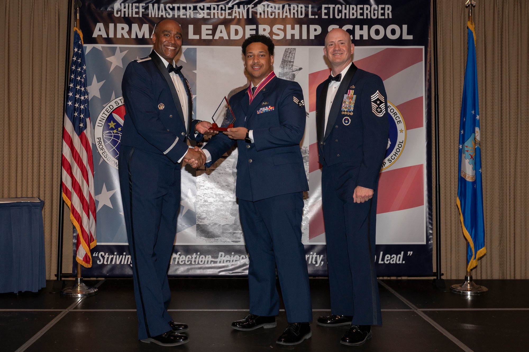 Three men stand on a stage together while the first and second man shake hands, while holding an award together.