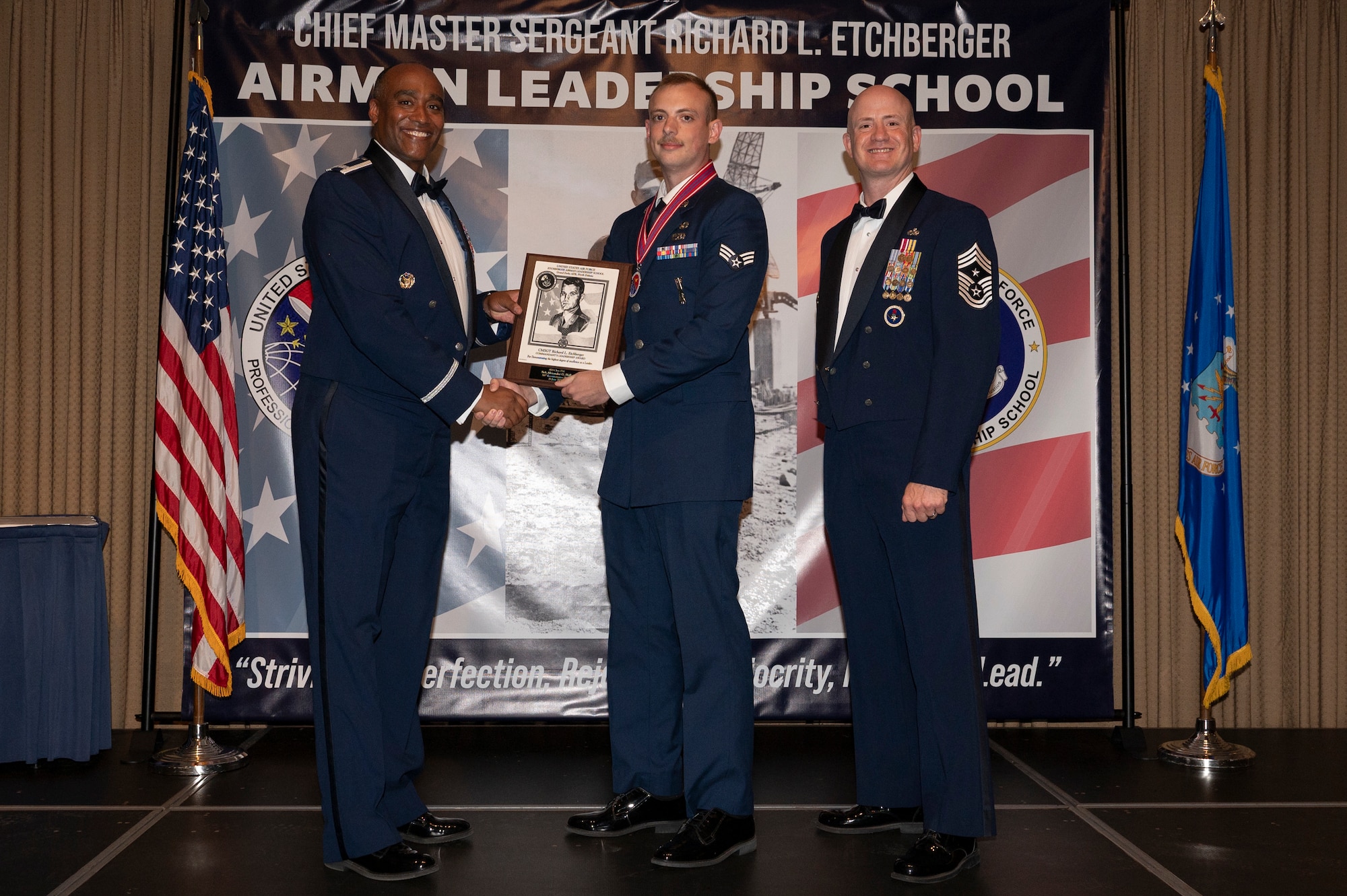 Three men stand on a stage together while the first and second man shake hands, while holding an award together.