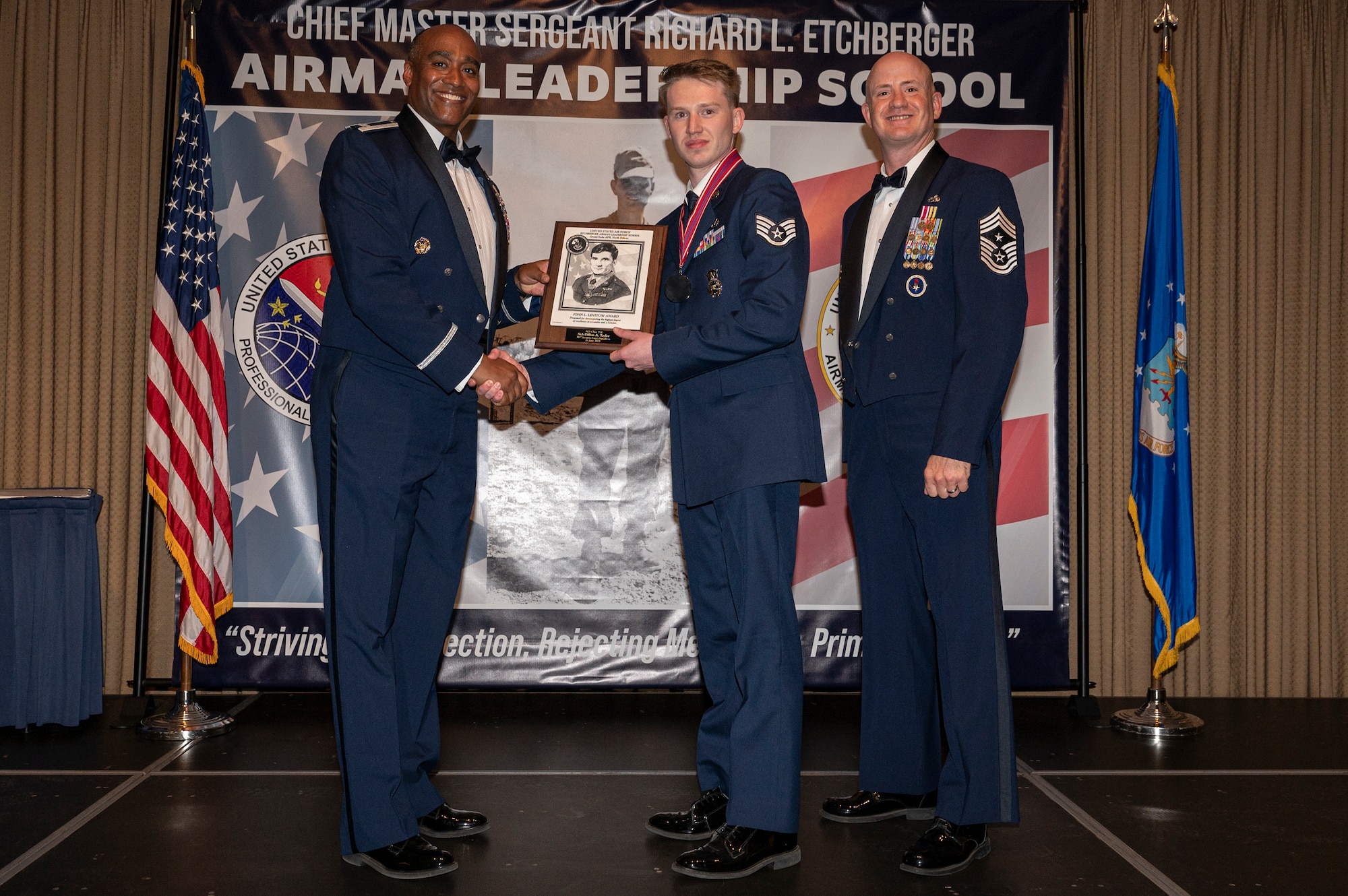 Three men stand on a stage, the first and the second shake hands while holding an award together.