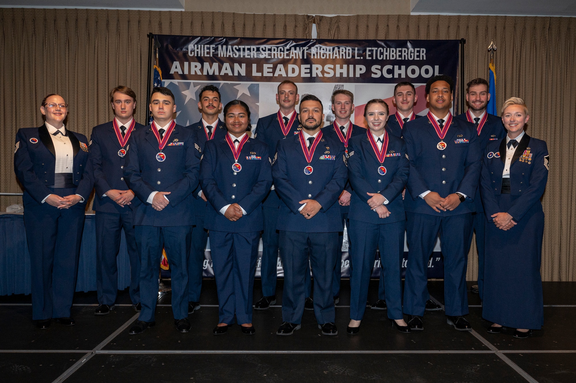 A group of students and their two supervisors stand on a stage for a group photo.