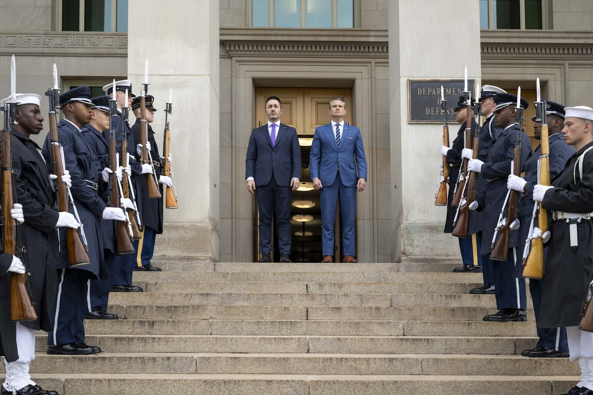 Two men wearing business attire stand atop stairs, as people wearing formal military uniforms stand in formation.