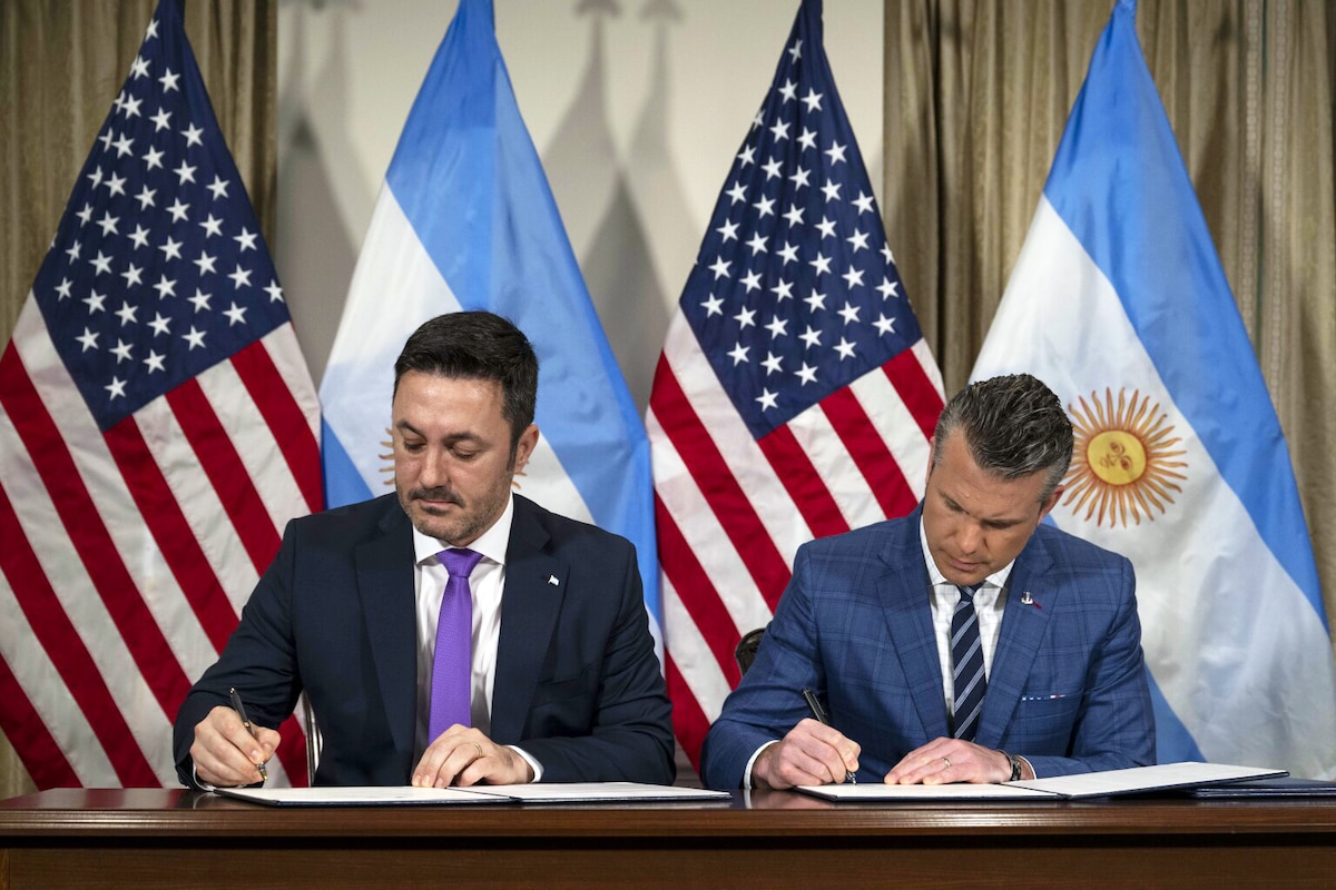 Two men in business attire sit at a desk and sign a paper. Behind them are the American and Argentine flags.