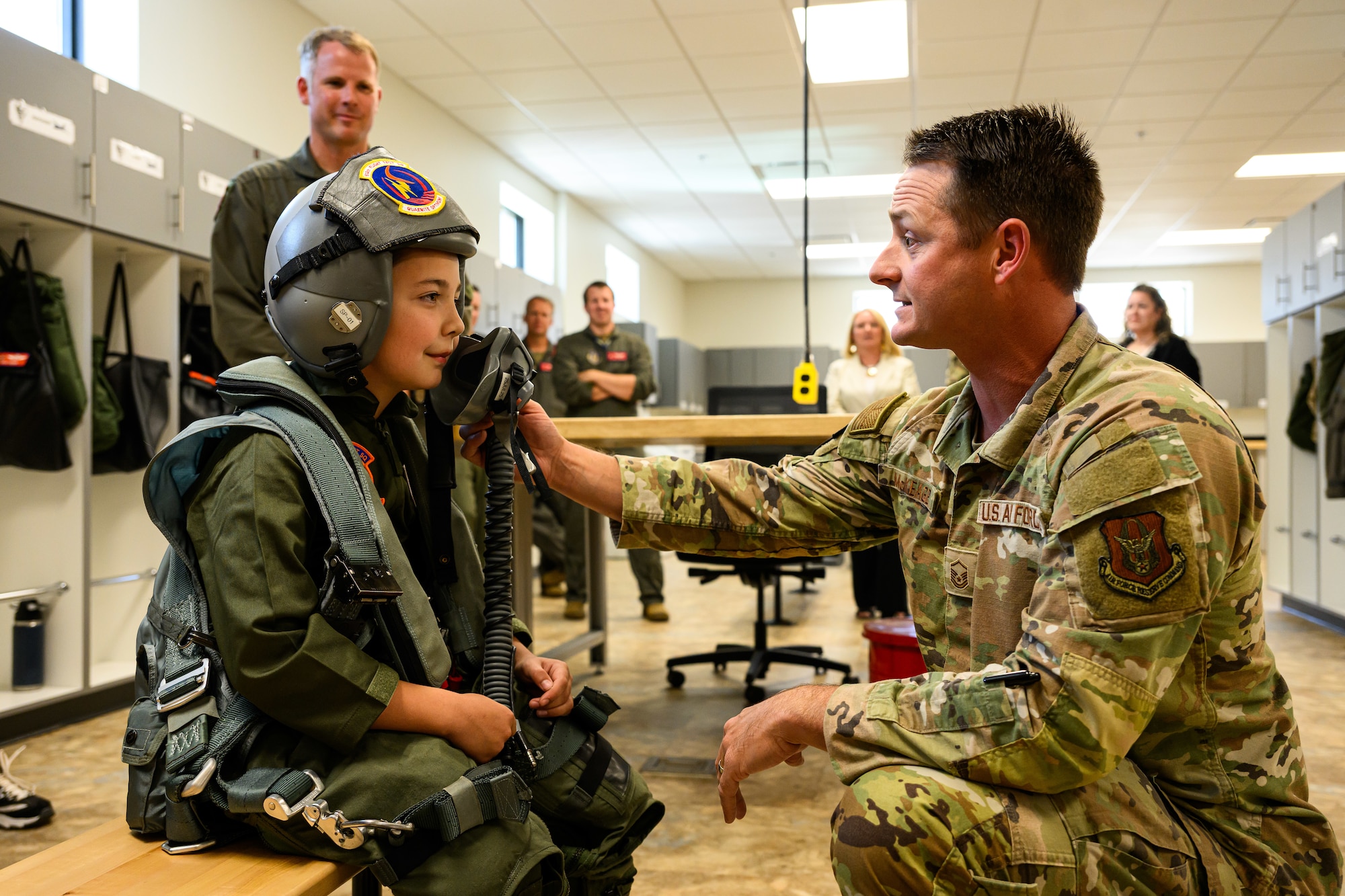 Dylan sits on a bench wearing flight equipment while an aircrew member at right holds the breathing mask to Dylan's face.