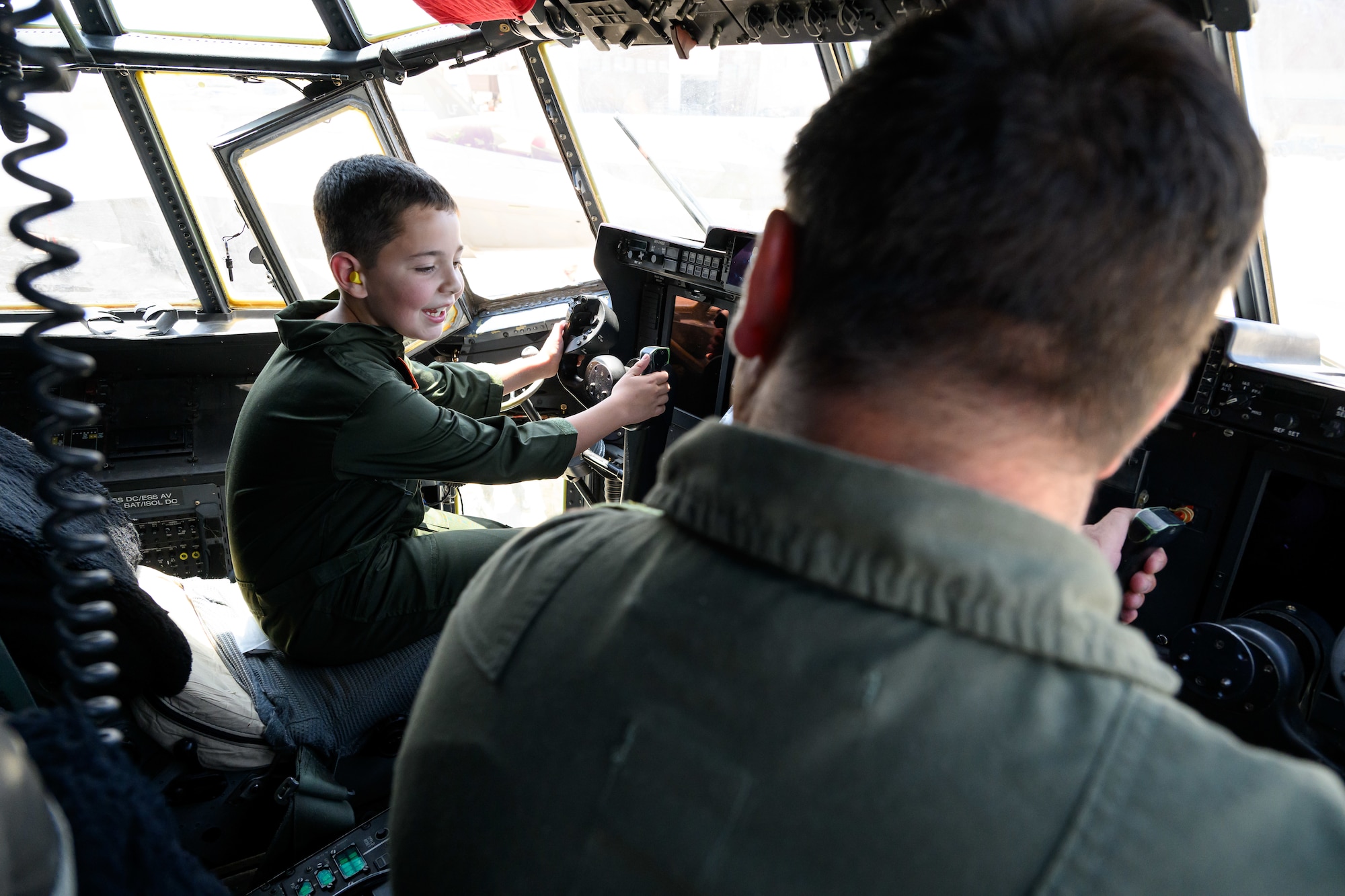 Dylan conners sits in the pilot seat of a C-130 while a pilot in the foreground talks to him.