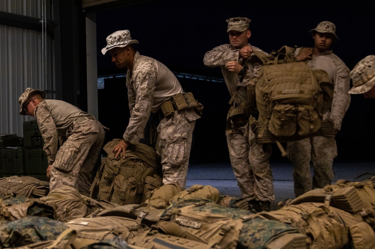 Four people wearing camouflage uniforms are inside, grabbing large backpacks off the floor.