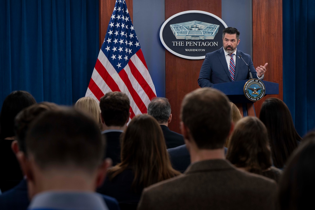 A man in civilian business attire is standing behind a podium in a room, speaking to a group of people with their backs to the camera. There is an American flag to his left and a sign on the wall behind him with a drawing of a five-sided building that reads, “The Pentagon.”