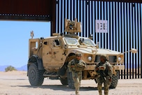 Two soldiers in combat gear walk away from a military vehicle on a sunny day in desert-like conditions, with a tall security barrier in the background.