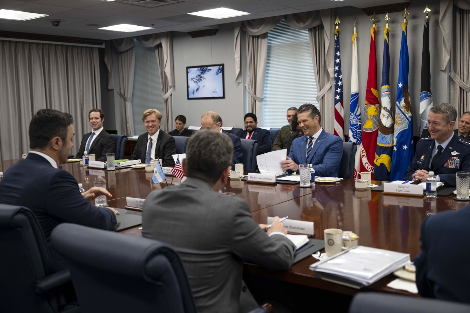 People wearing formal military uniforms and business attire smile while sitting around a wooden table with papers in front of them.