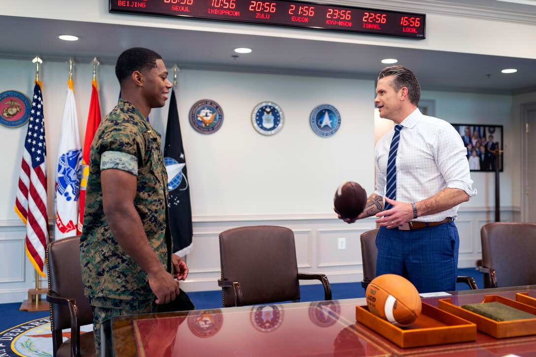 A man uniform chats with a man in a suit holding a football inside a room with flags and clocks in the background.