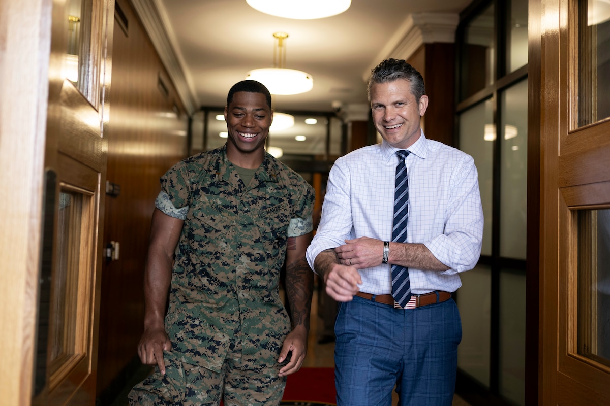 Defense Secretary Pete Hegseth and a Marine stand and smile next to each other in a hallway.