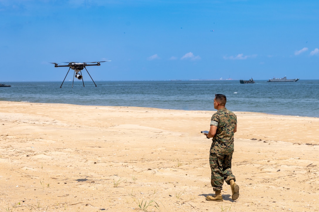 A Marine holding a remote control walks on a beach as a drone flies to the left while two ships sail to right under a blue sky.