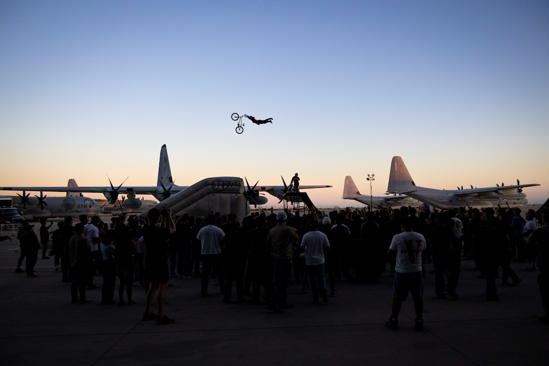 A biker performs a leg free trick over aircraft on a flight line as a crowd watches in silhouette with a pinkish horizon.