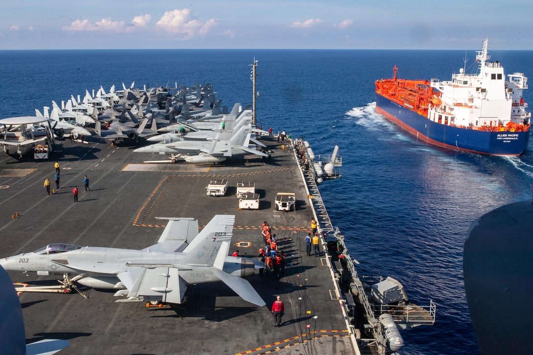 Sailors work near aircraft aboard a ship sailing toward a smaller red, white and blue ship during the day.